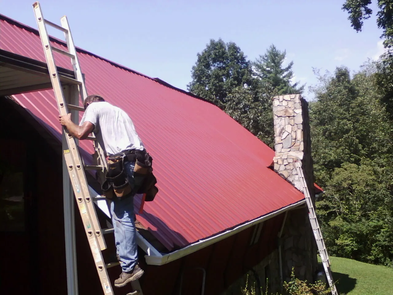 Roofer on a ladder beside a red metal roof and stone chimney — Soffit & Fascia Repair by Roofing Brothers in La Porte